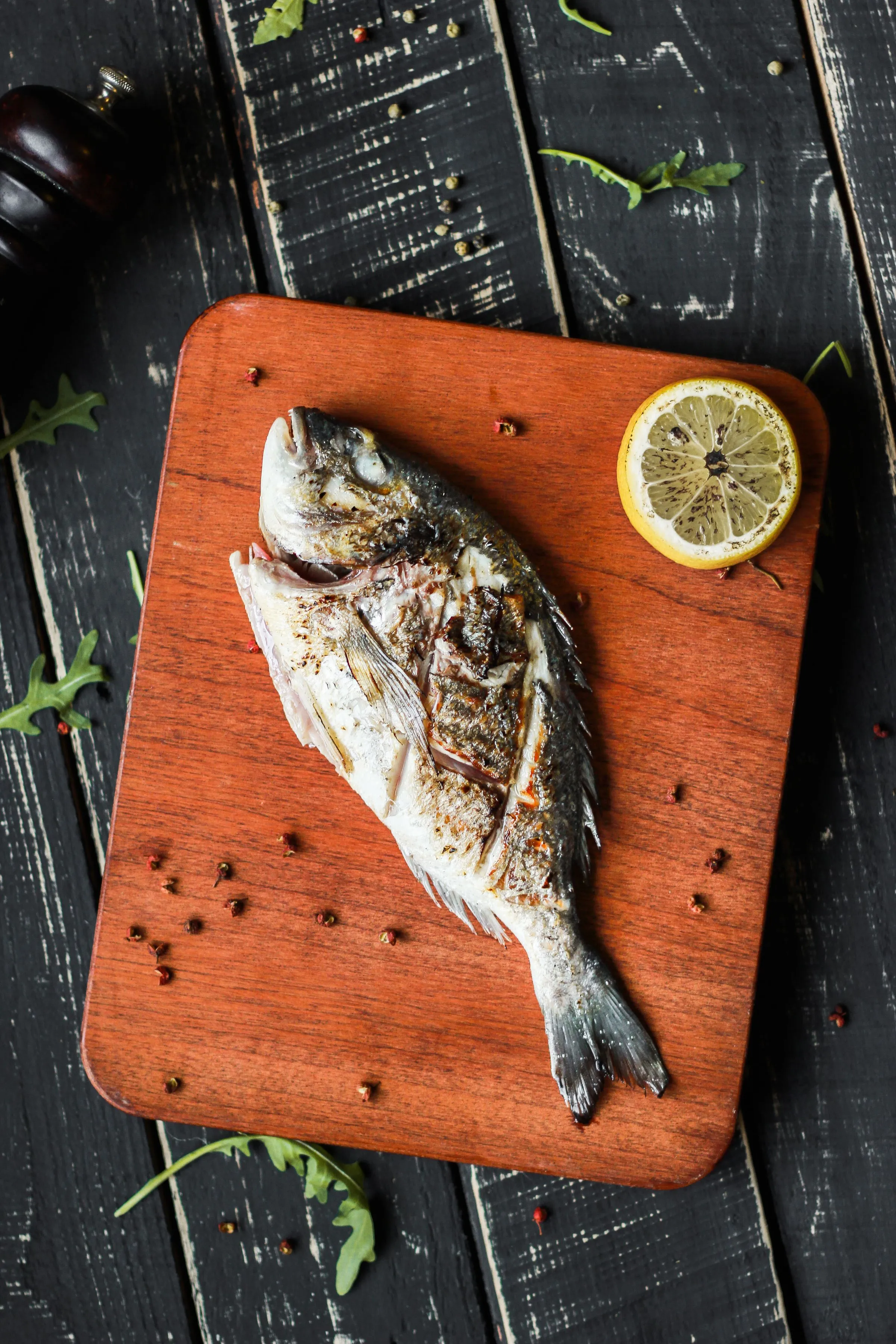 A whole grilled fish on a wooden board with lemon and pepper.